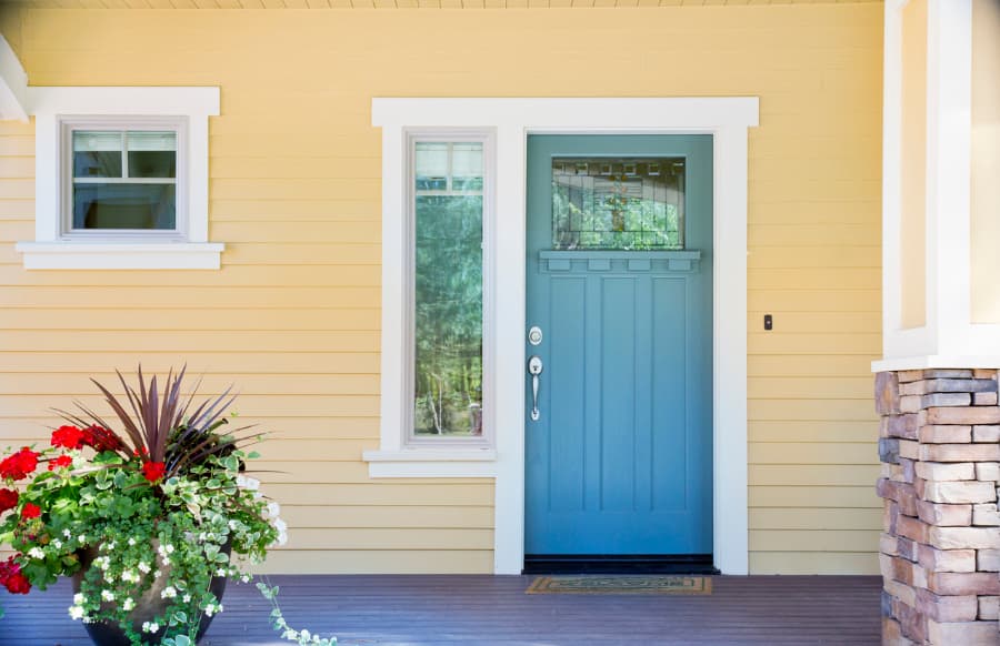 A Front Entrance Of A Home With A Blue Door, Yellow Siding, And A Flowerpot In Daytime